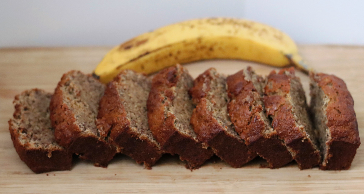 Un banana bread sans gluten tranché sur une planche de bois et une banane en arrière plan.