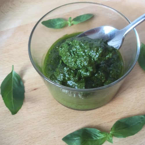 basil pesto in a glass jar and a spoonful being taken out of it. basil leaves around the jar to decorate