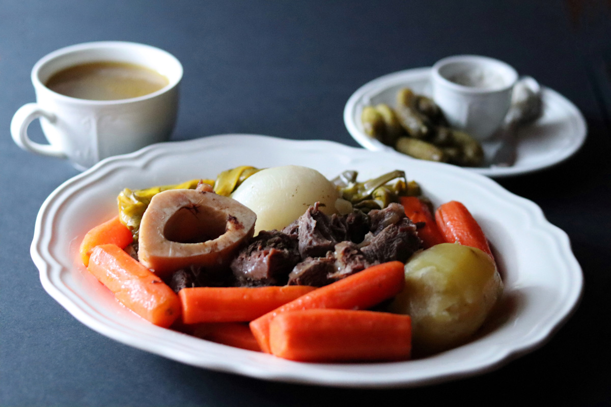 Beef stew in a large white plate and a cup of bone broth in the background with a plate of cornichons and coarse salt