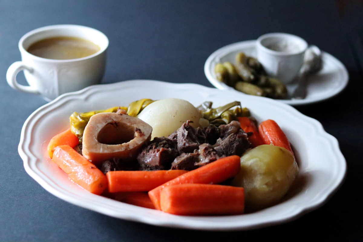 un pot au feu servi dans une grande assiette creuse blanche avec une tasse de bouillon, du gros sel et des cornichons