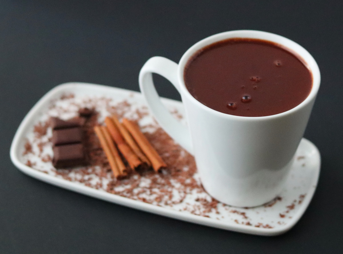 a white mug full of hot chocolate on a white plate with 4 cinnamon sticks and a chocolate bar and chocolate sprinkles to decorate the plate