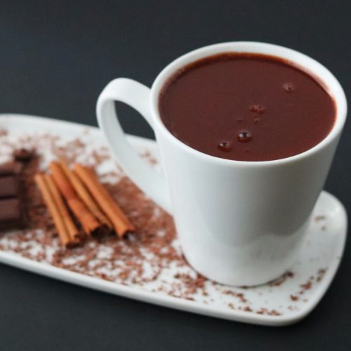 a cup of hot chocolate on a white plate with cinnamon sticks and a chocolate bar.