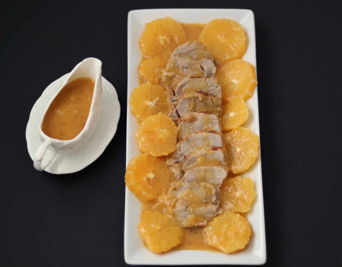 pork tenderloin on a serving plate with orange slices for garnish, a sauce boat full of orange sauce on the side
