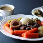 Beef stew in a large white plate and a cup of bone broth in the background with a plate of cornichons and coarse salt