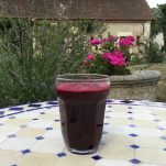 a glass of beetroot and carrot juice on a table in a garden with flowers and a house in the background
