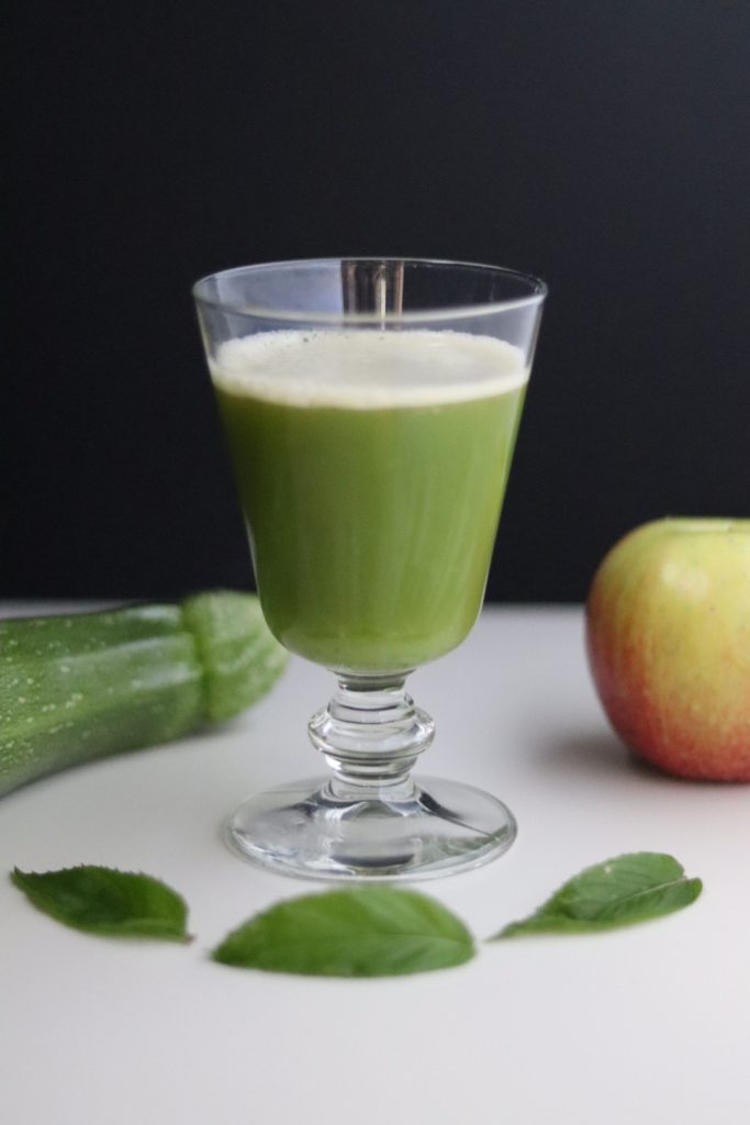 a glass of zucchini and apple juice, a zucchini, an apple and 3 mint leaves around the glass