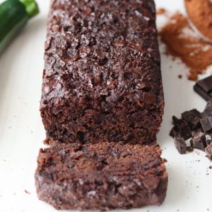 un gâteau chocolat courgette avec une courgette sur le côté, une cuillerée de cacao en poudre et des morceaux de chocolat