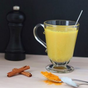 a glass cup full of golden milk surrounded by 2 cinnamon sticks, a spoon full of turmeric, a spoon full of coconut oil and a pepper mill