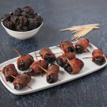 A bunch of bacon wrapped prunes on a white dish and a bowl full of prunes and toothpicks in the background