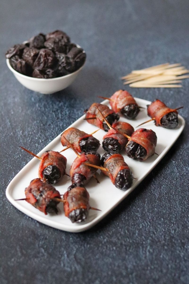 A bunch of bacon wrapped prunes on a white dish and a bowl full of prunes and toothpicks in the background