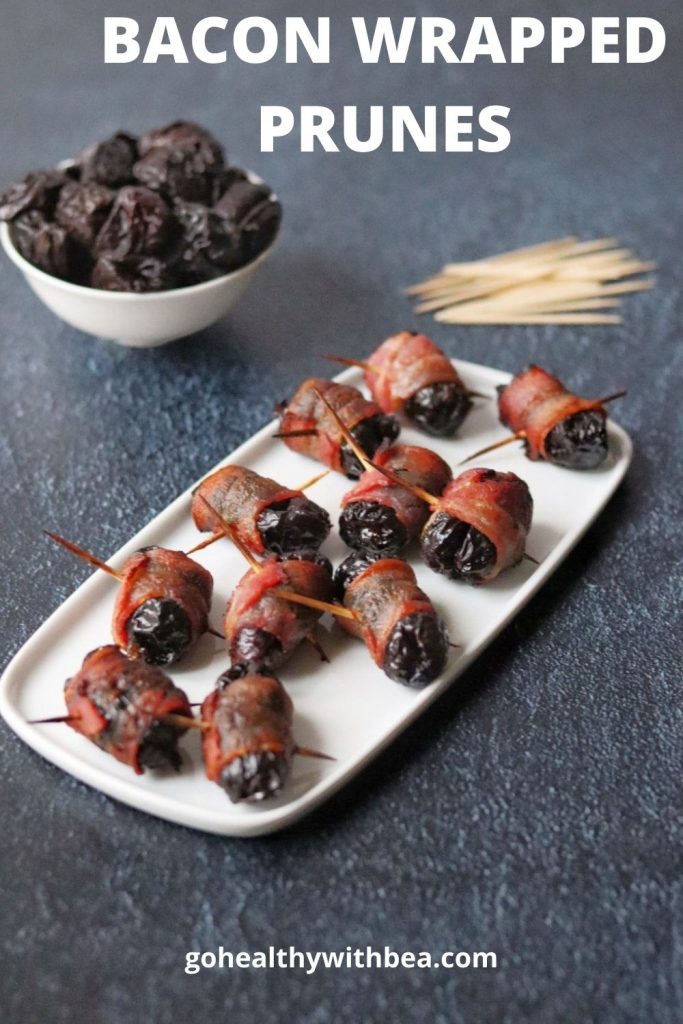 A bunch of bacon wrapped prunes on a white dish and a bowl full of prunes and toothpicks in the background