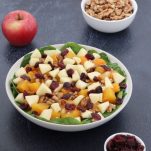 fall harvest salad in a white plate, an apple, a bowl of walnuts and a small bowl of dried cranberries