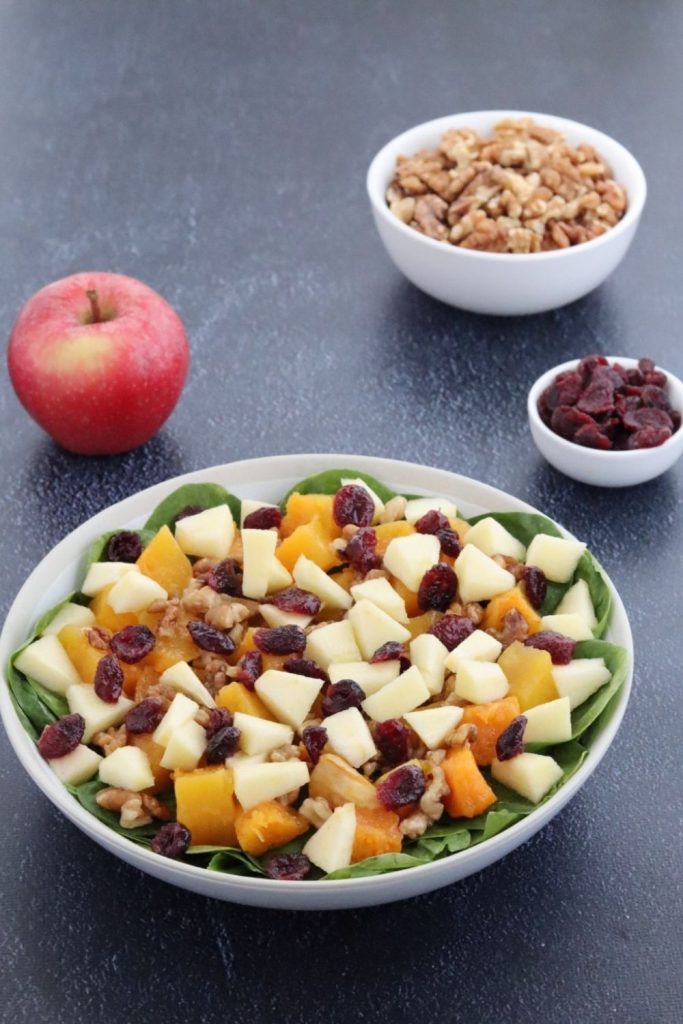 fall harvest salad in a white plate, an apple, a bowl of walnuts and a small bowl of dried cranberries