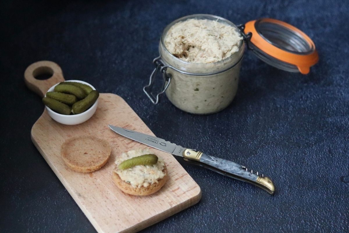a glass jar full of chicken rillettes next to a wooden board with bread rolls and cornichons on it