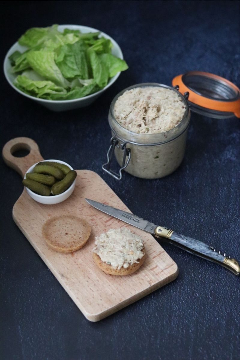 a glass jar full of chicken rillettes next to a wooden board with bread rolls and cornichons on it and a bowl of lettuce in the background