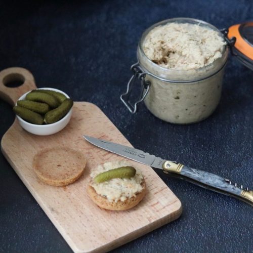 a glass jar full of chicken rillettes next to a wooden board with bread rolls and cornichons on it