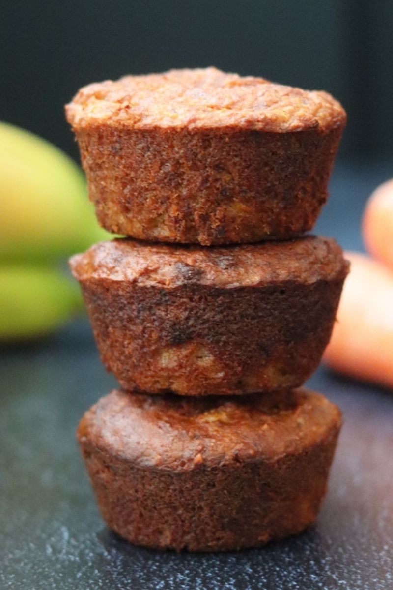 3 banana carrot muffins piled up with bananas and carrots in the background 