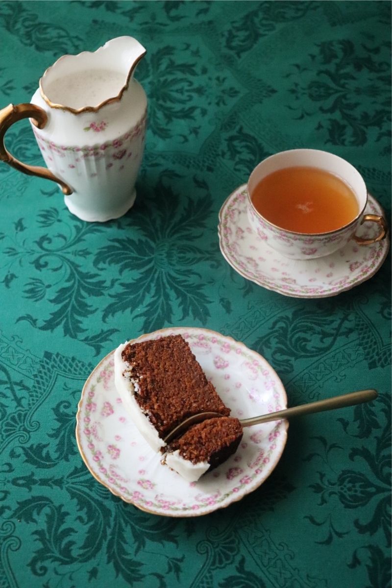 a slice of gingerbread loaf on a plate with a fork cutting a piece, a cup of tea and a pot in the background