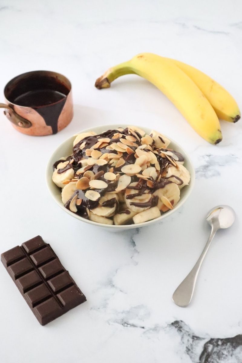 a bowl full of banana dessert with chocolate a small saucepan filled with chocolate in the background with 2 bananas, a chocolate bar and a spoon in the foreground