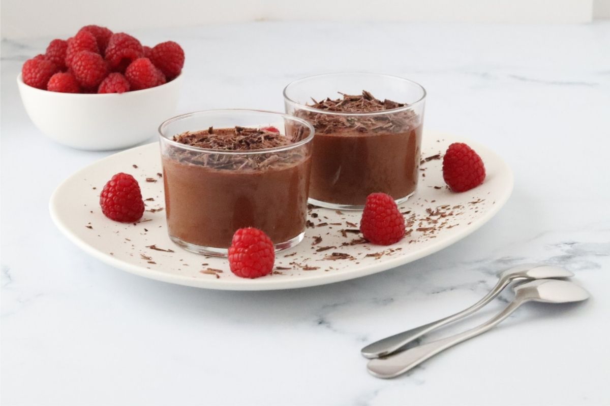 2 glasses full of chocolate mousse on a white plate with raspberries and small pieces of chocolate on the plate to decorate. A white bowl full of raspberries in the background and 2 teaspoons next to the plate, in the foreground