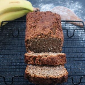 a banana bread with 2 pieces sliced on a baking rack bananas and a wooden spoon in the background