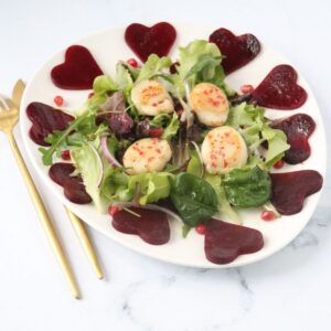 une assiette blanche sur laquelle on peut voir de la salade mesclun avec de l'oignon rouge, des graines de grenade et 4 noix de saint jacques parsemées de baies roses moulues et des coeurs de betterave tout autour de l'assiette