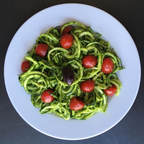 a white plate full of zucchini noodles coated with pesto and topped with cherry tomatoes and a black olive
