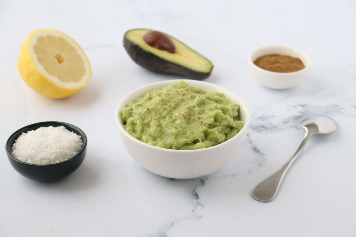 a white bowl full of guacamole, a spoon on the side, sea salt ground cumin half an avocado and half a lemon in the background