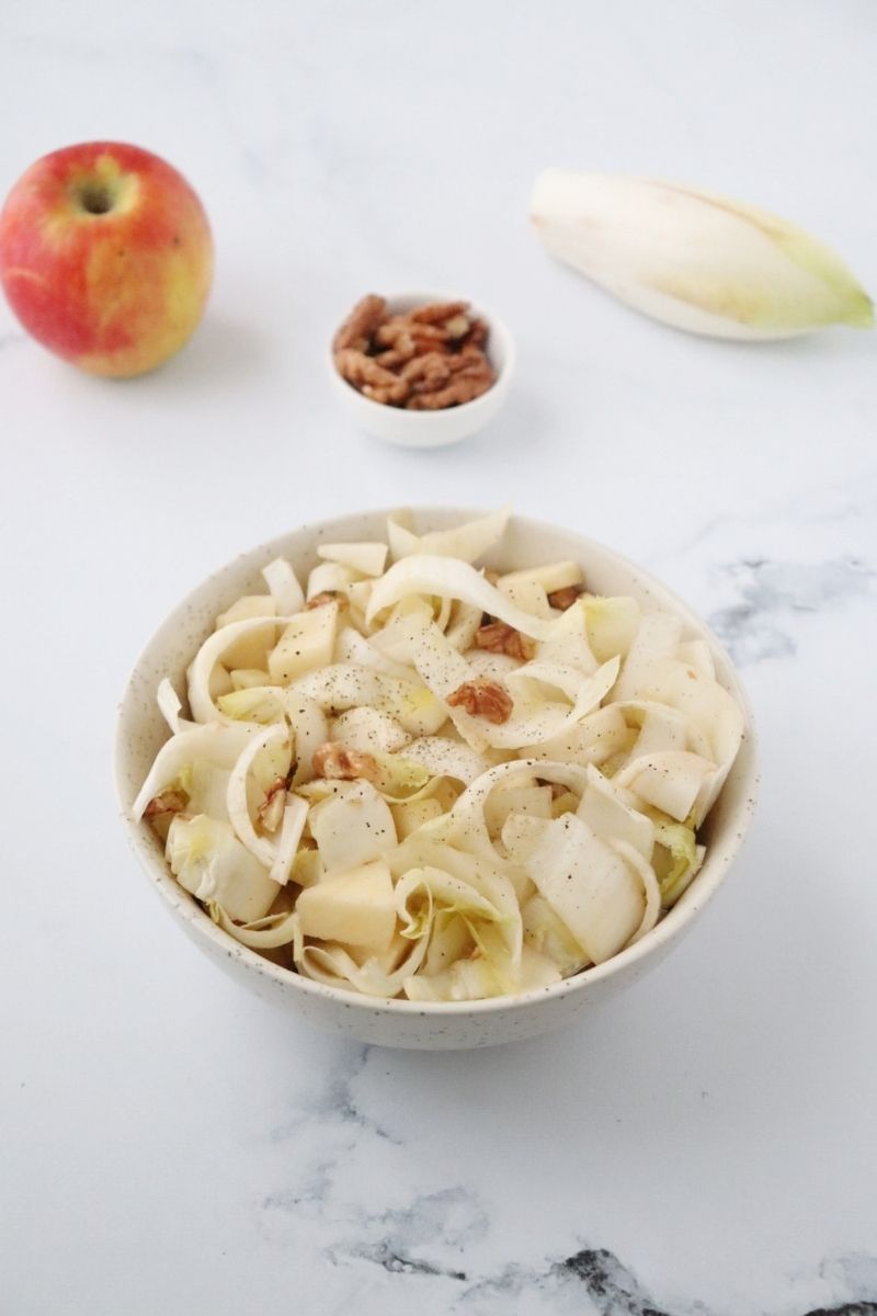 un saladier rempli de salade d'endives aux noix et aux pommes, une pommes, un mini bol rempli de noix et une endive à côté du saladier