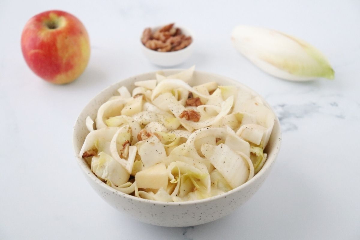 un saladier rempli de salade d'endives aux noix et aux pommes, une pommes, un mini bol rempli de noix et une endive à côté du saladier