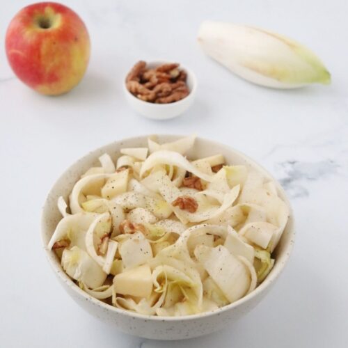a bowl full of endive, walnut and apple salad, next to it there is an apple, a small bowl full of walnuts and an endive