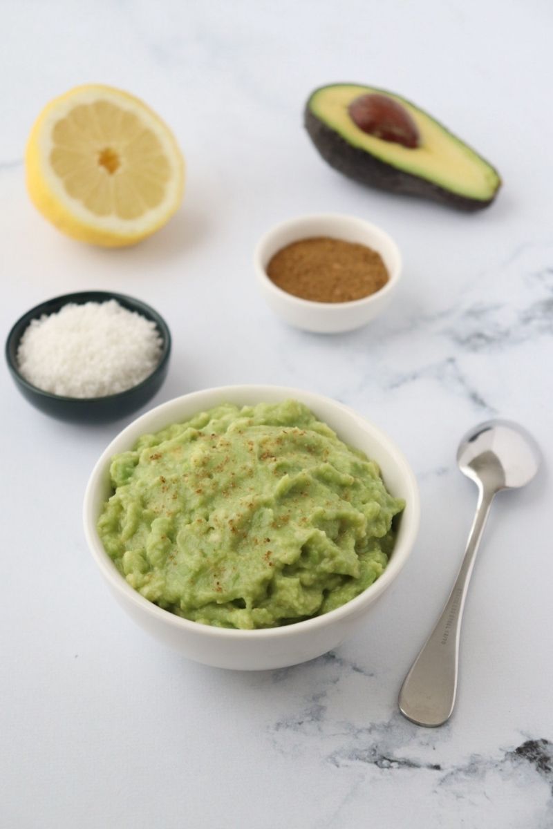 un bol rempli de guacamole, une cuillère, du sel du cumin un demi citron et un demi avocat tout autour du bol