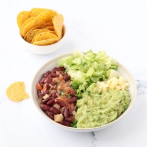 a bowl filled with guacamole, lettuce, cheese and kidney beans and some corn tortilla chips on the side