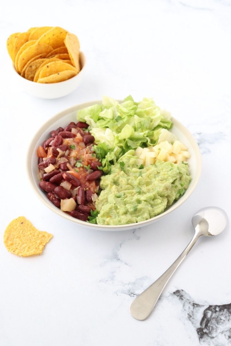 a bowl filled with guacamole, lettuce, cheese and kidney beans and some corn tortilla chips on the side.