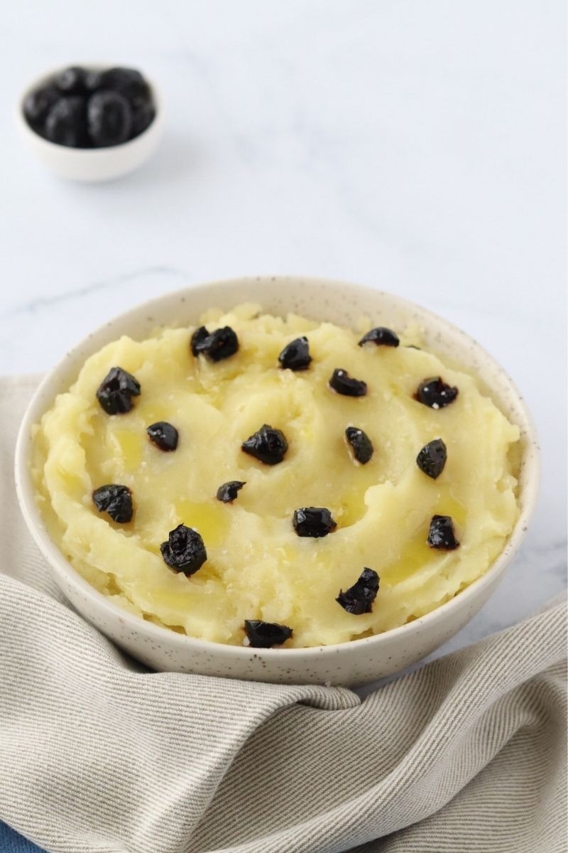 a bowl full of mashed potatoes topped with pieces of black olives, a small bowl of black olives in the background