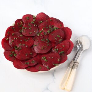a plate full of beetroot carpaccio with chopped parsley sprinkled on top and serving spoons on the side of the plate