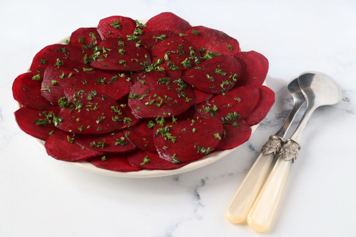 a plate full of beetroot carpaccio with chopped parsley sprinkled on top and serving spoons on the side of the plate