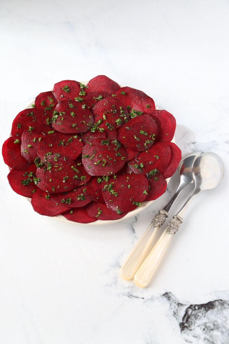 a plate full of beetroot carpaccio with chopped parsley sprinkled on top and serving spoons on the side of the plate