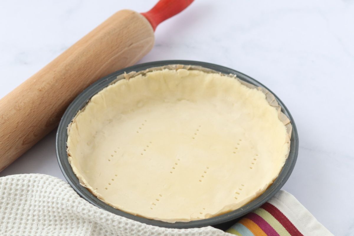 A pie crust in a pie tin with a table cloth and a rolling pin on the side.