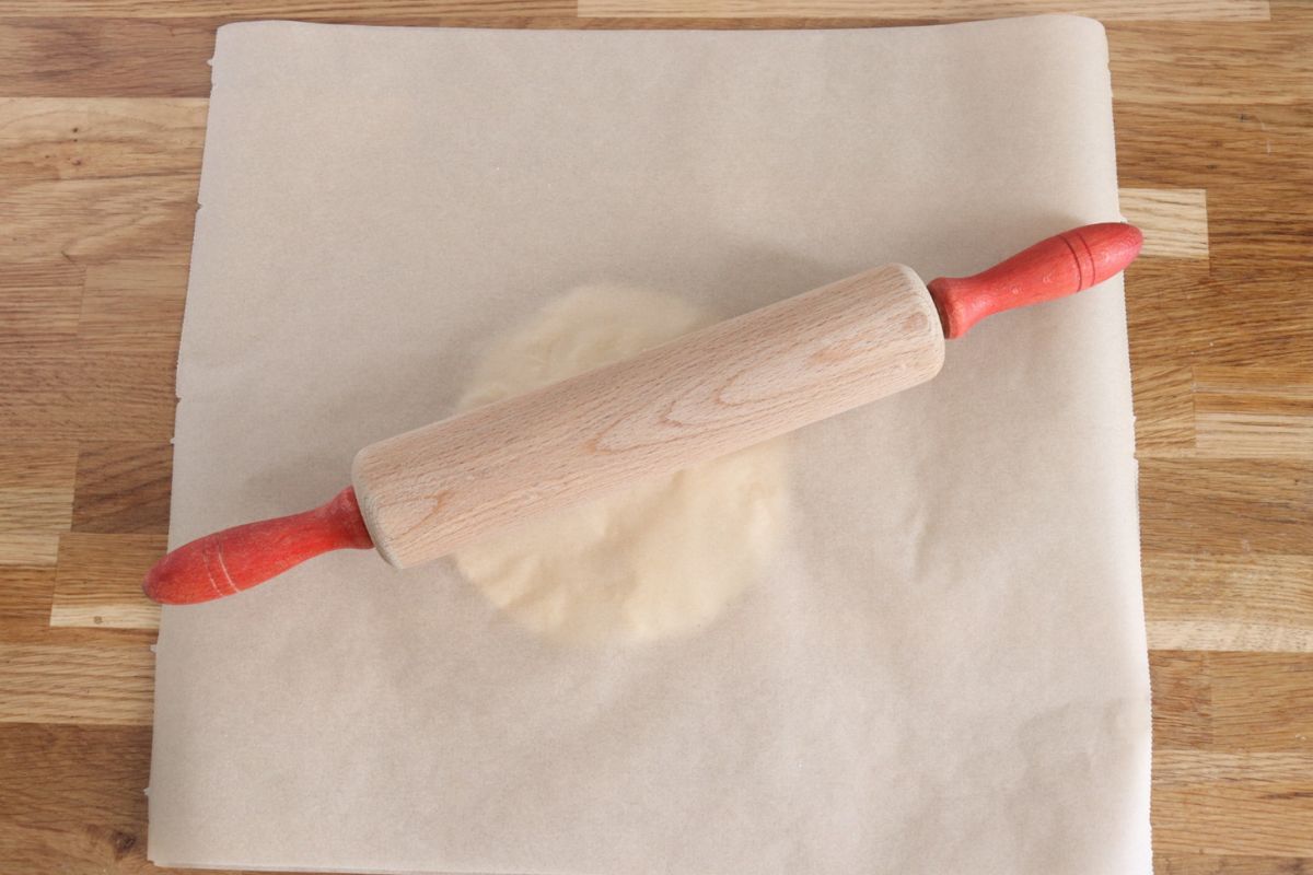 A flattened dough between 2 parchment papers a rolling pin over it.