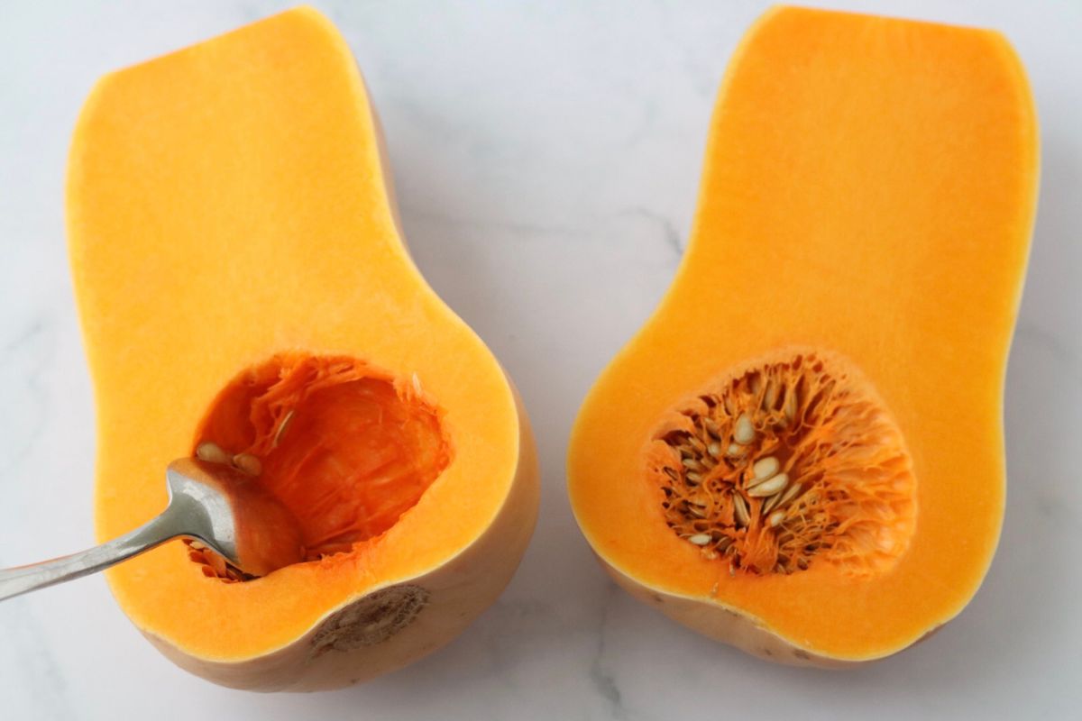 a spoon removing the seeds of one of the halves of a butternut squash