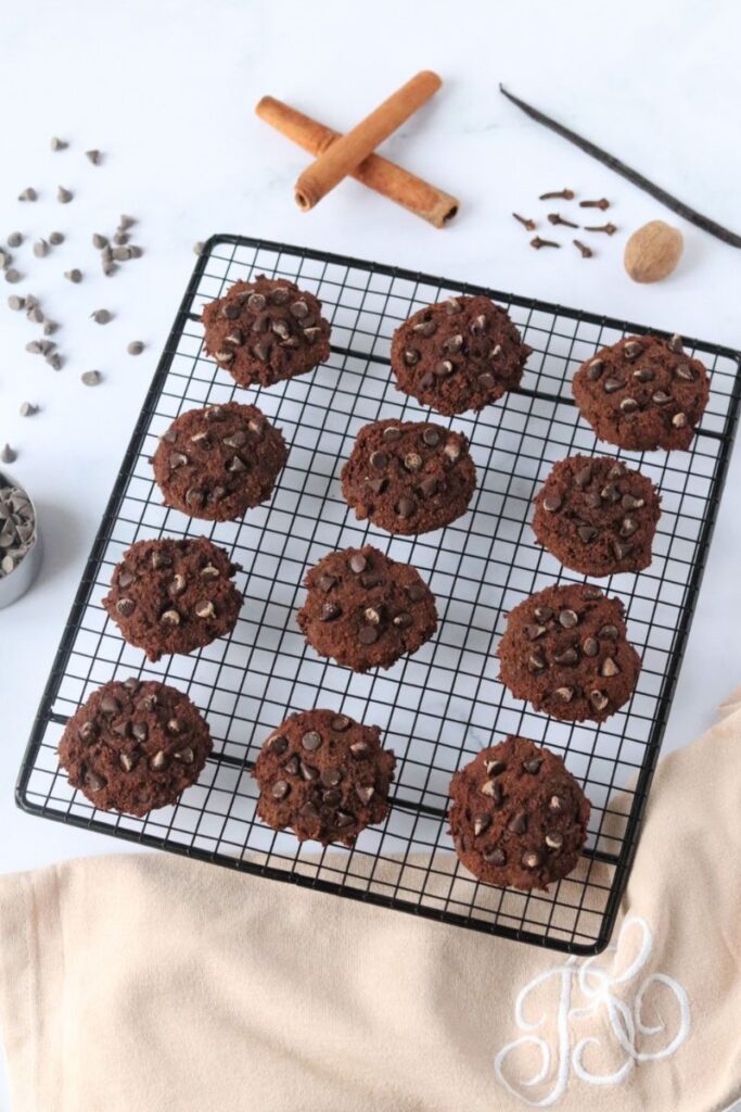 Des cookies à la farine de souchet sur une grille à patisserie avec des pépites de chocolat, des bâtons de cannelle, de la vanille, des clous de girofle et une noix de muscade tout autour.