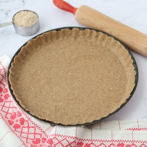 a pie crust in a pie tin with a table cloth and a rolling pin, a table cloth and a cup full of flour on the side