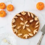 a pumpkin pie decorated with crust leaves, mini pumpkins around it with a plate, a fork and a cake server
