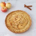 an apple tart a glass plate, 3 apples and 2 cinnamon sticks on the side