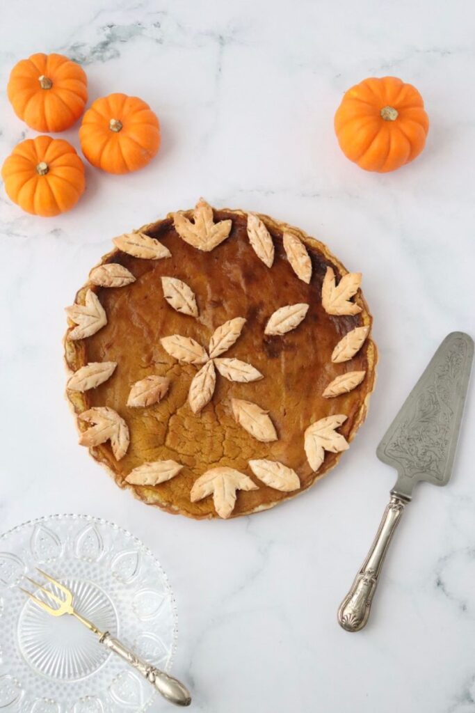 a pumpkin pie decorated with crust leaves, mini pumpkins around it with a plate, a fork and a cake server