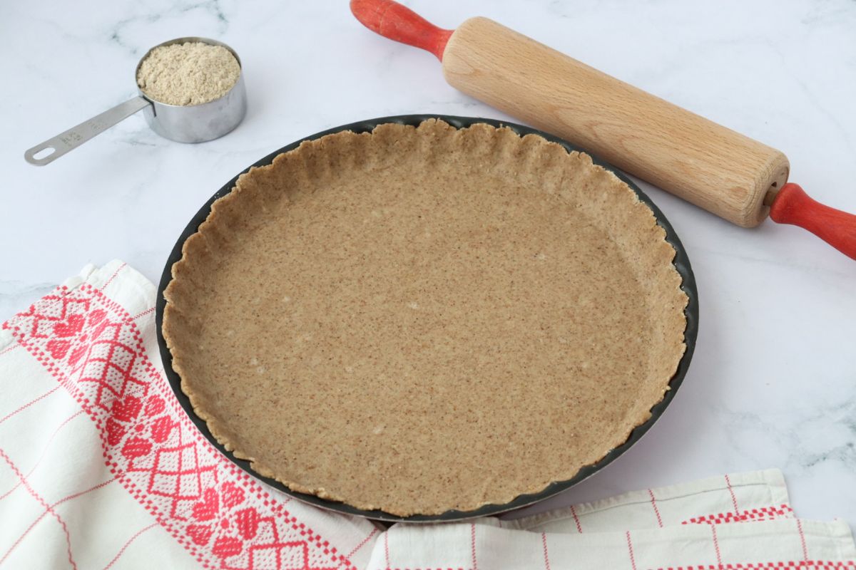 a pie crust in a pie tin with a table cloth and a rolling pin, a table cloth and a cup full of flour on the side