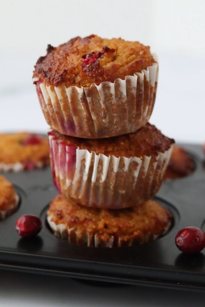 3 orange cranberry muffins piled together in a muffin pan, fresh cranberries placed on the pan to decorate