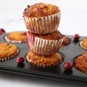 3 orange cranberry muffins piled on top of each other on a muffin pan and fresh cranberries placed on the sides