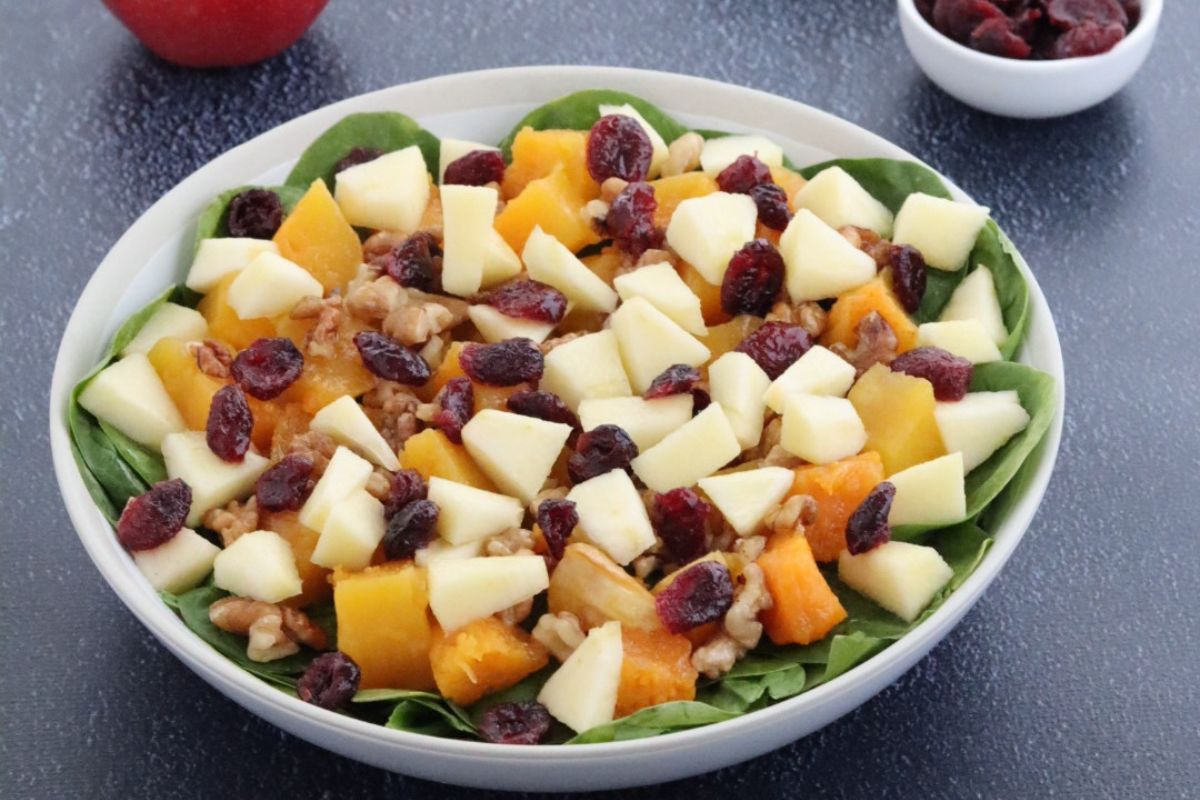 fall harvest salad in a white plate, an apple, a bowl of walnuts and a small bowl of dried cranberries